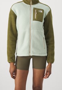 Fleecejacka i mintgrön och olivgrön, med dragkedja fram, bröstficka och textilstruktur; ihop med olivgröna shorts.