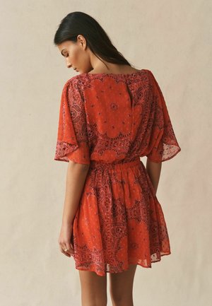 Hosszú sötét hajú nő laza, rövid piros ruhát visel lila paisley mintákkal, megköthető derékkal és széles ujjal, háttal áll.