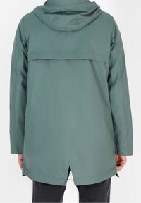 Tylny widok osoby w turkusowej wodoodpornej kurtce z kapturem, długimi rękawami i małym wentylacyjnym otworem z tyłu, w połączeniu z ciemnymi spodniami.