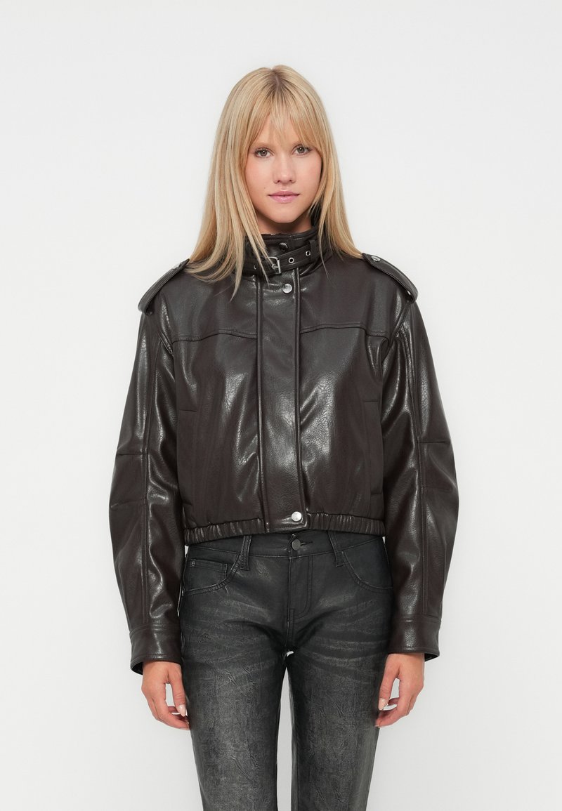 Blonde Frau, die eine dunkelbraune Lederjacke mit Schnallenkragen und schwarze strukturierte Hose trägt, steht vor einem weißen Hintergrund.