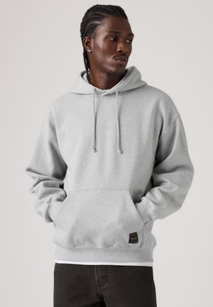 Schwarzer Mann mit Zöpfen, der einen hellgrauen Hoodie und schwarze Hose trägt, schaut nach links mit den Händen in der vorderen Tasche des Hoodies.