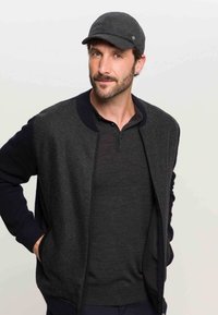 Dark grey jacket with navy ribbed cuffs and collar, zippered closure, layered over a dark grey polo shirt, paired with a matching flat cap.