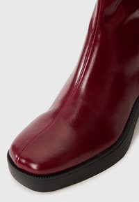 Burgundy leather ankle boot with a glossy finish, pointed toe, and a textured black rubber sole. Features vertical stitching details.