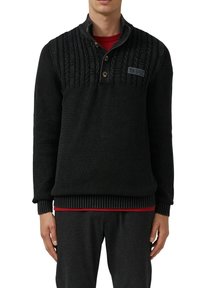 Mann trägt einen schwarzen strukturierten Strickpullover mit Knöpfen und einem roten Hemd darunter, kombiniert mit dunklen Hosen.