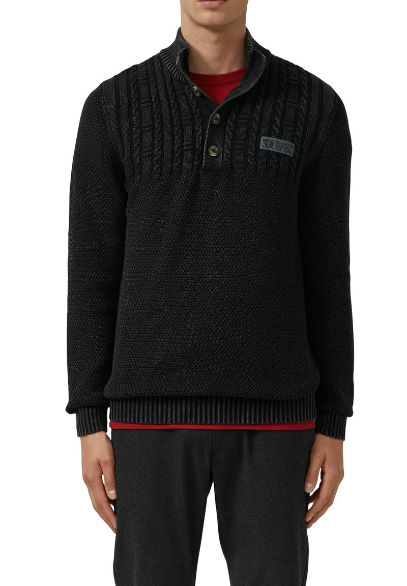 Mann trägt einen schwarzen strukturierten Strickpullover mit Knöpfen und einem roten Hemd darunter, kombiniert mit dunklen Hosen.