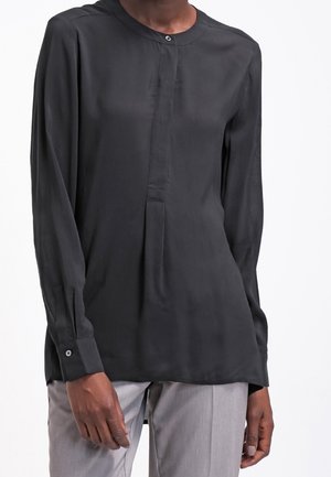 Person trägt eine langärmelige schwarze Bluse mit Stehkragen und graue Hose, steht vor einem weißen Hintergrund.