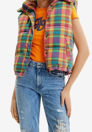 Person trägt eine mehrfarbige karierte gesteppte Weste über einem orangefarbenen T-Shirt mit Grafik und hoch taillierte, zerrissene blaue Jeans mit Strassdetails.