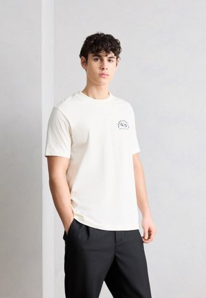 Mladý muž s kučeravými tmavými vlasmi, oblečený v bielom tričku s krátkym rukávom a malým logom na hrudi a čiernych nohaviciach, stojaci s jednou rukou vo vrecku.