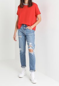 Joven vistiendo una camiseta roja de manga corta, jeans claros rasgados, calcetines blancos y zapatillas blancas, de pie con una mano en el bolsillo.