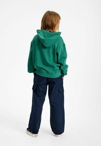 Sudadera verde con capucha y un ajuste relajado, combinada con pantalones cargo azul marino con bolsillos laterales y zapatillas blancas con detalles rojos.