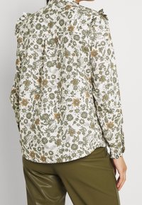 Chemise à imprimé floral avec des fleurs vertes et orange sur fond blanc, tissu texturé et détails de volants aux épaules.