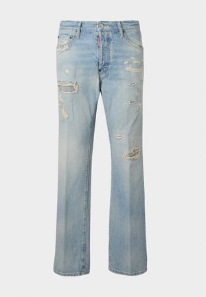 Jeune homme portant un sweat crème, un jean bleu clair déchiré et des bottes rouge foncé avec des lacets rouges, debout devant un fond blanc uni.