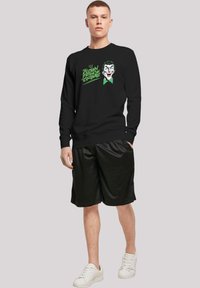 Svart hettegenser med en tegneseriefigur i grønt og hvitt, kombinert med svarte shorts og hvite sneakers. Glatt stofftekstur.