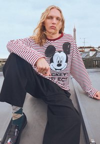 Tricou cu mâneci lungi în dungi roșii și albe, cu un mare grafic Mickey Mouse, asortat cu pantaloni negri largi și șosete cu model.