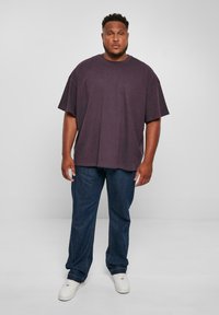 Tricou din bumbac violet supradimensionat cu mâneci scurte, purtat cu blugi albaștri și adidași albi, stând în fața unui fundal simplu.