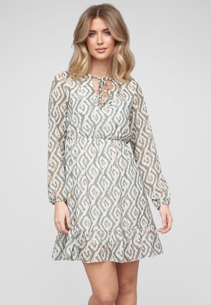 Jeune femme aux cheveux blonds mi-longs portant une robe longue à manches longues, à motifs, longueur genoux, avec un col à nouer et un ourlet volanté.