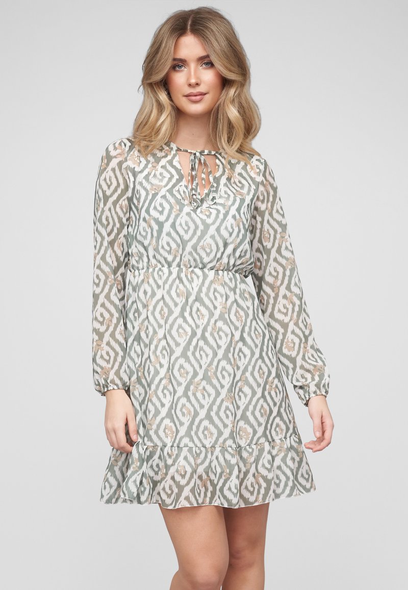 Jeune femme aux cheveux blonds mi-longs portant une robe longue à manches longues, à motifs, longueur genoux, avec un col à nouer et un ourlet volanté.