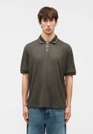 Νέος άνδρας που φοράει πόλο πουκάμισο ελιάς με λευκή περιμετρική ραφή στο γιακά και μπλε τζιν, στέκεται μπροστά σε απλό φόντο.