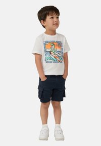Witte katoenen T-shirt met een kleurrijke dinosaurustekening, gecombineerd met marineblauwe cargoshorts en witte sneakers, met een luxe textuur.