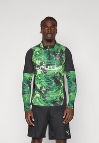 Grøn camouflage langærmet skjorte med sorte accenter, halv-lynlås krave og logo detaljer på brystet; parret med sorte shorts.