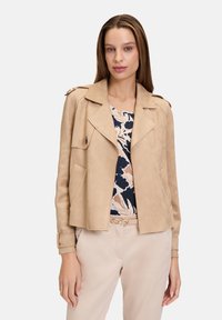 Veste en daim beige clair au design cropped, avec des revers notés et des poches latérales. Portée par-dessus un haut à imprimé floral et un pantalon de couleur claire.