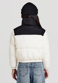 Steppjacke mit einem cremeweißen Körper und einem schwarzen oberen Abschnitt. Verfügt über elastische Bündchen und Saum. Getragen zu hellblauen Jeans.