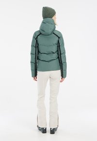 Chaqueta de plumas aislante verde con un diseño ajustado, capucha, detalles en negro y mangas largas. Combinada con pantalones beige claros y botas de esquí.