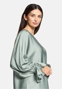 Haut en satin vert clair avec une coupe décontractée, des manches larges resserrées aux poignets et une texture lisse et brillante.