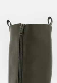 Acercamiento del eje de bota de cuero verde oscuro con un zipper vertical y dos lazos en la parte superior sobre un fondo blanco.