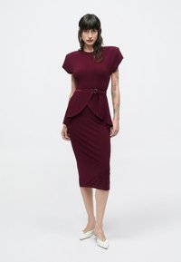 Bordowa sukienka o dopasowanym kroju, z krótkimi rękawami, warstwowym fasonem peplum oraz paskiem podkreślającym talię. Gładka faktura tkaniny.