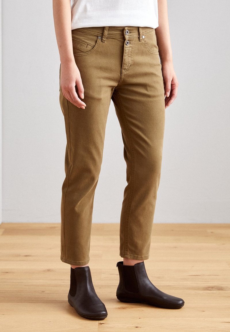 Personne portant un pantalon marron ajusté et des bottines noires, debout sur un sol en bois clair contre un mur gris uni.