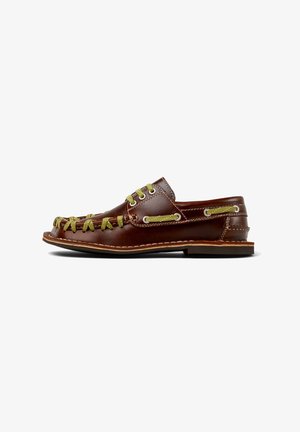 Zapato slip-on de cuero marrón con cordones y costuras verdes, con punta redondeada, ojales laterales y suela plana.