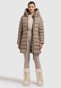 Beige Pufferjacke mit Kapuze, versehen mit horizontaler Steppung, Frontreißverschluss und Seitentaschen. Begleitet von einem hellbeigen, figurbetonten Outfit und weißen Schneestiefeln.