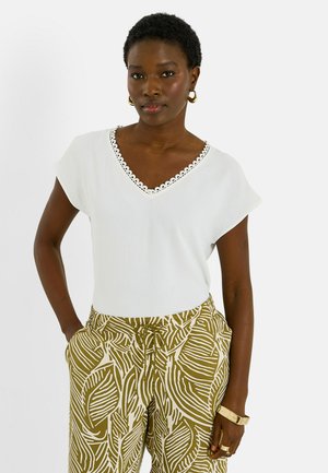 Mujer con top blanco de escote en V con borde festoneado, pantalones verde oliva con estampado de hojas blancas, pendientes de aro dorados y brazalete y anillos dorados.
