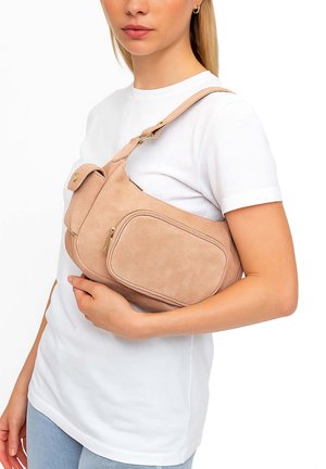 Vrouw met een beige suède crossbodytas met een voorvak met rits en een klepzak met knoop, gekleed in een wit T-shirt en lichtblauwe spijkerbroek.