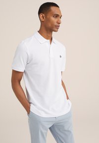 Camisa polo blanca de un tejido transpirable, con cuello clásico, mangas cortas y un logo negro en el pecho. Combinada con pantalones azul claro.