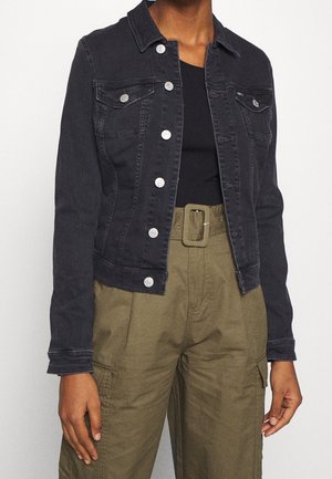 Personne portant une veste en jean noire à boutons sur un haut noir et un pantalon taille haute vert olive avec une grande ceinture et des poches latérales.