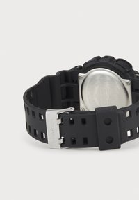Black G-Shock watch featuring a textured rubber band with cutout patterns, silver metal clasp, and a large circular case with a stainless steel back.