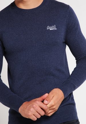 Homme portant un pull à manches longues bleu marine avec un logo blanc SuperDry sur la poitrine gauche, mains jointes à la taille.