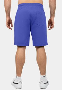 Shorts di cotone viola con cintura elastica e tasca posteriore, caratterizzati da un taglio dritto e da una texture liscia, mostrati da dietro.