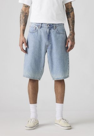 Férfi világoskék farmer rövidnadrágban, fehér bokazokniban, fehér sportcipőben és fehér pólóban, karjai lazán az oldala mellett áll.