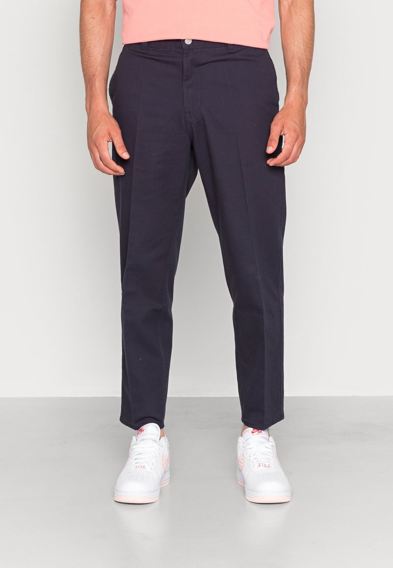 Marineblaue Chinos mit lockerer Passform, ausgestattet mit zwei Fronttaschen, einem Knopfverschluss und einer glatten Baumwolltextur. Kombiniert mit weißen Sneakern.