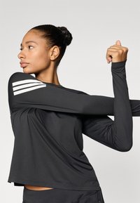 Μαύρο αθλητικό μπλουζάκι με μακριά μανίκια και άνετη εφαρμογή, με τρεις λευκές ρίγες στον αριστερό ώμο και λεία, ελαφριά υφάσματα.