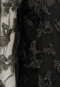 Tissu noir avec des broderies argentées complexes en motifs floraux, comportant des sections transparentes et des détails de surface texturés.