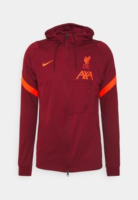 Maronenfarbene Kapuzenjacke mit orangefarbenen Akzenten, ausgestattet mit einem Reißverschluss, einer Tasche und dem auffällig platzierten Liverpool FC-Logo. Hergestellt aus glattem Stoff.