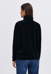 Zwarte corduroy pullover met een hoge kraag en ribbeltextuur, gecombineerd met donkere spijkerjeans. Het ontwerp heeft lange mouwen en een comfortabele pasvorm.