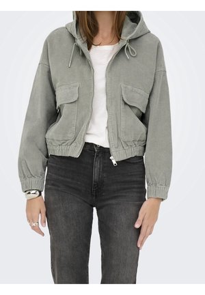 Kvinde iført lys grå cordfløjl jakke med lynlås og hætte, hvid t-shirt og mørkegrå jeans, med synligt armbånd og ringe på håndleddet.