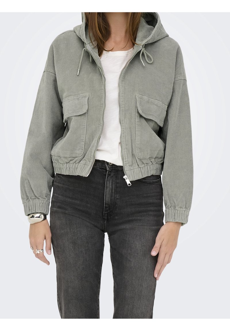 Femme portant une veste zippée en velours côtelé gris clair avec capuche, un t-shirt blanc et un jean gris foncé, avec un bracelet et des bagues visibles au poignet.