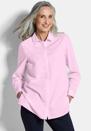 Glimlachende vrouw van middelbare leeftijd met grijs haar, gekleed in een lichtroze blouse met knopen en donkere broek, handen in de zakken, staand tegen een witte achtergrond.