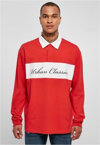Polo roșu cu mânecă lungă, decorat cu o bandă orizontală albă pe care este inscripționat textul "Urban Classics", guler clasic și țesătură moale din bumbac.